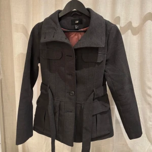 Grå kappa från H&M - Snygg grå kappa från H&M med knappar och bälte i midjan. Jackan har långa ärmar och praktiska fickor framtill.