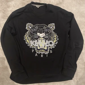 Svart sweatshirt från Kenzo - Säljer en svart sweatshirt från Kenzo med ett stort broderat tigerhuvud och texten 'Kenzo Paris' på framsidan. Tröjan har långa ärmar och en rund halsringning. Perfekt för en stilren och trendig look.