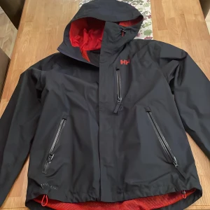 Svart vindjacka från Helly Hansen - Säljer en svart vindjacka från Helly Hansen med röd insida. Jackan har en justerbar huva och flera praktiska fickor med dragkedja. Perfekt för blåsiga dagar och utomhusaktiviteter.