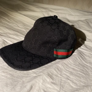 Svart keps från Gucci - Snygg svart keps från Gucci med det ikoniska GG-mönstret. Kepsen har en justerbar rem baktill och en grön och röd randdetalj på sidan. Perfekt för att ge din outfit en lyxig touch.