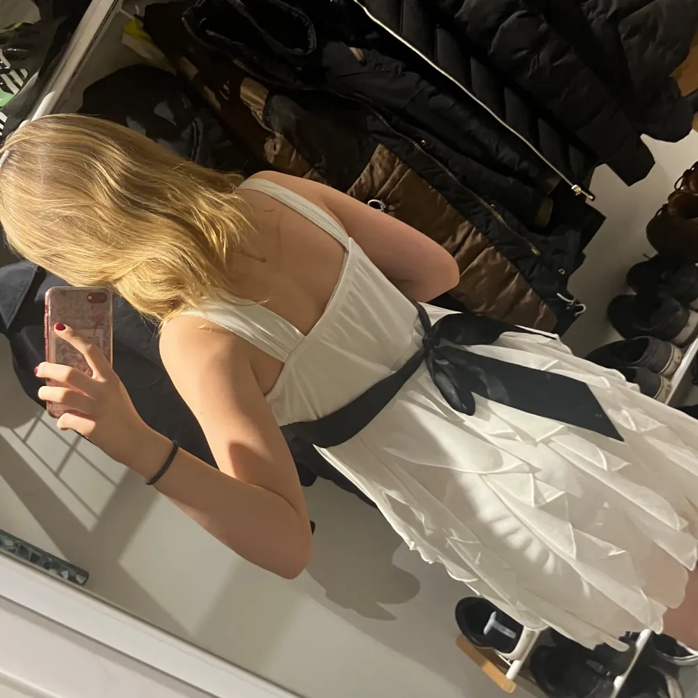 Elegant vit klänning med ett svart band i midjan och en dekorativ rosett baktill. Klänningen har en omlott-design med volanger och är ärmlös, vilket ger en luftig och stilren look.. Mekot.