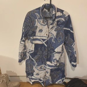 Mönstrad kappa i blått och vitt - Säljer en unik mönstrad kappa i blått och vitt med paisley-inspirerat mönster. Kappan har långa ärmar och knappar framtill. Perfekt för att ge din outfit en bohemisk touch.