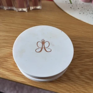 Highlighter i vit dosa - Elegant highlighter i en vit dosa med spegel. Produkten har en skimrande champagnefärg och är perfekt för att ge ansiktet en strålande lyster. Dosan har en stilren design med en logotyp i guld på locket.