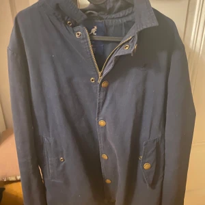 Mörkblå jacka från Kangol - Säljer en stilren mörkblå jacka från Kangol med dragkedja och knappar. Jackan har en klassisk krage och två fickor framtill. Insidan är fodrad med ett rutigt mönster i bruna och beige toner.