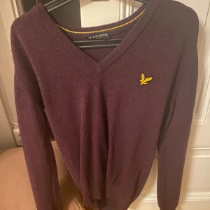 Vinröd tröja från Lyle & Scott - Snygg vinröd tröja från Lyle & Scott med deras ikoniska gula logga på bröstet. Tröjan har en v-ringad hals och långa ärmar, perfekt för kyligare dagar. Den är stilren och passar bra till både jeans och chinos.