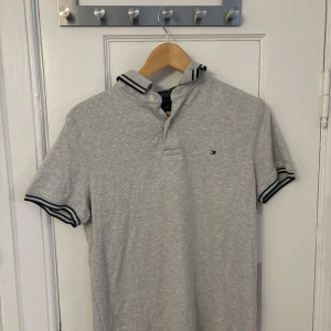 Grå pikétröja från Tommy Hilfiger - Säljer en stilren grå pikétröja från Tommy Hilfiger med klassisk krage och knappar. Tröjan har svarta och vita ränder på kragen och ärmsluten samt en liten logotyp på bröstet. Perfekt för en avslappnad men snygg look.