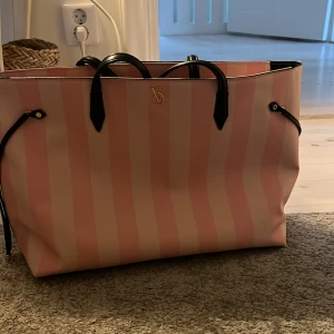 Randig shoppingväska med svarta handtag - Snygg shoppingväska med rosa och beige ränder och svarta handtag. Väskan har en öppen design med en rymlig insida, perfekt för shoppingturer. Men även perfekt för skolan jag fick platts med alla mina mappar i den. Den har även en liten logga framtill.