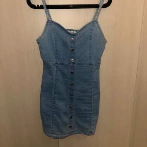 Jeansklänning med knappar - Trendig jeansklänning med knappar framtill och justerbara axelband. Perfekt för en avslappnad stil.