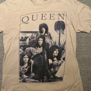 Beige Queen t-shirt - Säljer en beige t-shirt med ett stort tryck av bandet Queen på framsidan. T-shirten är från Queen Official Merch