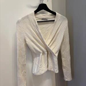 Vit omlottopp från Berskha - Snygg vit omlottopp med ribbad struktur. Toppen har långa ärmar och en elegant omlottdesign som ger en smickrande passform. Perfekt för både vardag och festliga tillfällen.