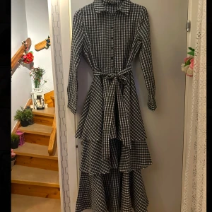 Svartvit rutig klänning med volanger - Snygg svartvit rutig klänning med långa ärmar och knappar framtill. Klänningen har volanger och ett knytbälte i midjan för en smickrande passform. Perfekt för en stilren look.