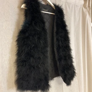 Svart pälsväst från Nardaler - Passar S/M om man vill ha en tjockare tröja under. Snygg svart pälsväst från Nardaler, perfekt för att ge din outfit en lyxig touch. Västen har en quiltad insida och är så fluffig och fin då den är gjord på bra material. Strutsfjädrar.