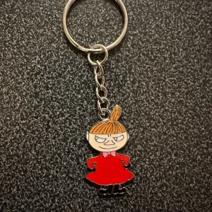 Nyckelring med Lilla My - Charmig nyckelring med motiv av Lilla My från Mumintrollen. Figuren är klädd i en röd klänning och har sitt karakteristiska uttryck. Perfekt för att ge dina nycklar en personlig touch!