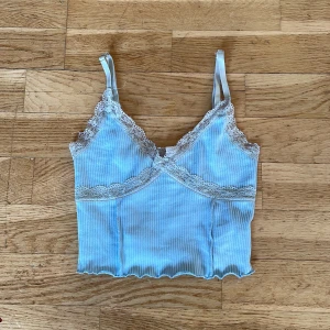 Ljusblå ribbad linnetopp från Divided - Säljer en söt ljusblå ribbad linnetopp från Divided med spetsdetaljer längs kanterna. Toppen har smala axelband och en croppad passform. Perfekt för varma dagar eller som ett lager under en tröja.