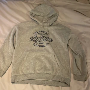 Grå hoodie med vintage-tryck - Säljer en grå hoodie med tryck 'The Original Vintage Clothing Co.' på framsidan. Den har en klassisk känguruficka och justerbar huva med dragsko. Perfekt för en avslappnad stil.