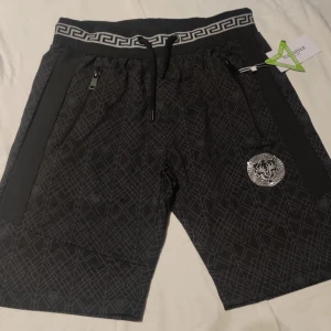 Svarta mönstrade shorts med dragsko nytt - Snygga svarta shorts med geometriskt mönster och en cool logga på benet. De har en elastisk midja med grekisk nyckel-design och dragsko för perfekt passform. Praktiska dragkedjefickor på sidorna.