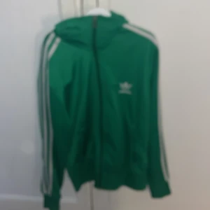Grön huvtröja från Adidas - Snygg grön huvtröja från Adidas med vita ränder längs ärmarna och en stor logga på ryggen. Tröjan har dragkedja och långa ärmar, perfekt för en sportig look.