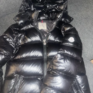  Moncler Maya - Snygg svart pufferjacka från Moncler med glansig finish och huva. Jackan har en dragkedja framtill och Moncler-logga på ärmen. Perfekt för kyliga dagar. Storlek M, bra skick.