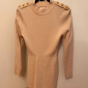 Beige ribbad klänning med guldknappar - Elegant beige ribbad klänning med långa ärmar och dekorativa guldknappar på axlarna. Perfekt för en stilren look. Från Raglady by tara Göteborg! Klänningen passar olika storlekar då den kramar om kroppen men även töjbar material! Aldrig använd! 