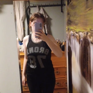 Svart ärmlös topp från BikBok - Säljer en svart ärmlös topp från BikBok med texten 'BRON 16' i grått framtill. Toppen är tillverkad i 100% polyester och har en sportig look med vita detaljer vid ärmhålen och halsringningen.