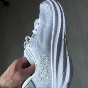 Vita Hoka One One Clifton 9 sneakers - Säljer ett par vita Hoka One One Clifton 9 sneakers. Skorna har en stilren design med snörning och en bekväm sula för optimal dämpning. Perfekta för löpning och vardagsbruk.