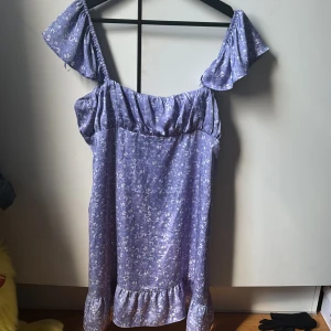 Lila blommig klänning från Zara - Charmig lila klänning från Zara med blommigt mönster. Den har offshoulder-ärmar och en volangdetalj vid fållen. Perfekt för en somrig look!