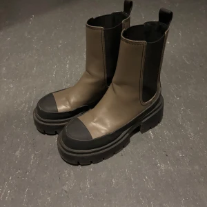 Khaki boots med svarta detaljer - Snygga bruna boots med svarta detaljer och grov sula. De har en elastisk panel på sidan för enkel på- och avtagning. Perfekta för en stilren look.