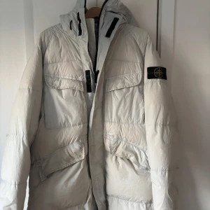 Vit dunjacka från Stone Island - Säljer en stilren vit dunjacka från Stone Island med huva och dragkedja. Jackan har flera praktiska fickor och den ikoniska loggan på ärmen. Perfekt för kalla dagar. Modellen är V0061 och går nypris för 14 000 kr. Mitt pris 7500. Köpt på spiga 3 Göteborg, har bara används en vinter, ett år gammal. 