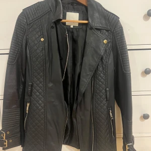 Svart skinnjacka från River Island - Snygg svart skinnjacka från River Island med quiltade detaljer och dragkedjor. Jackan har långa ärmar och stiliga guldfärgade knappar. Perfekt för en cool look!