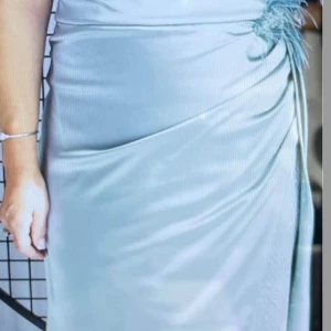 Ljusblå långklänning med glittriga detaljer - Elegant ljusblå långklänning med en v-ringad topp och glittriga detaljer. Klänningen har en draperad design med en slits och dekorativa fjädrar vid midjan. Perfekt för speciella tillfällen. Köpt från Cecilia Mode för 5000 kr endast använd 1 gång, priset kan diskuteras vid snabb köp