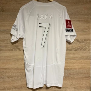 FA Cup Arsenal Saka XL - Snygg vit Arsenal fotbollströja från Adidas med Saka och nummer 7 på ryggen. Tröjan har korta ärmar och tryck på ärmarna med Emirates FA Cup och 'No room for racism'. Perfekt för fans av Arsenal och Saka! XL