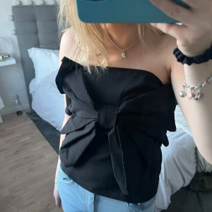 Svart axelbandslös topp med rosett - Snygg svart axelbandslös topp med en stor rosett framtill. Perfekt för en kväll ute eller en speciell tillställning. Toppen har en elegant och stilren design som passar till många olika outfits.