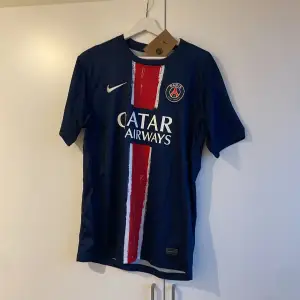 Säljer en blå PSG fotbollströja från Nike med röd och vit rand på framsidan. Tröjan har korta ärmar och klubbens emblem på bröstet. Perfekt för fotbollsfans!