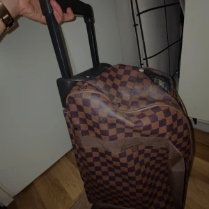 Brun rutig resväska - Snygg brun rutig resväska med dragkedja och teleskophandtag. Perfekt för resor med både handtag och hjul för enkel transport. Väskan har ett klassiskt rutmönster och är rymlig nog för alla dina behov.