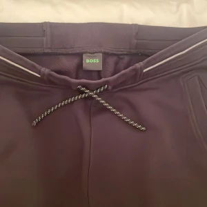 Svarta mjukisbyxor från Boss - Snygga svarta mjukisbyxor från Boss med snörning i midjan och en bakficka med logotyp. Perfekta för en avslappnad stil. Hela tracksuiten för 1200, koftan är storlek S och byxorna storlek M.