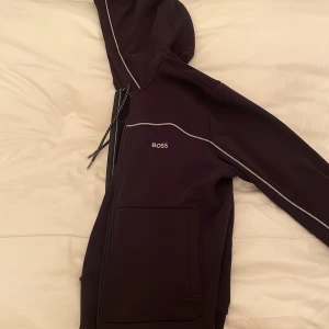 Svart hoodie från Boss - Snygg svart hoodie från Boss med dragkedja och huva. Den har vita detaljer längs ärmarna och snören vid huvan. Perfekt för en stilren och bekväm look.
