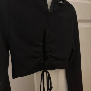 Svart långärmad tröja från hm - Snygg svart långärmad tröja från hm med knytning framtill och en elegant slits. Perfekt för en stilren look. Klänningen har en figurnära passform och är tillverkad i ett mjukt material.