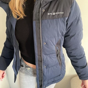 Sail Racing Jacka - Säljer en snygg blå dunjacka från Sail Racing med svart luva och axelparti. Jackan har dragkedja och praktiska fickor. Perfekt för kyliga dagar! inga defekter