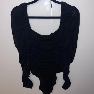 Svart långärmad topp - Snygg svart långärmad body  med rynkade detaljer och djup urringning. Perfekt för en stilren look. För fler bilder hör av er.