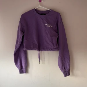 Lila croppad tröja från Bershka - Säljer en snygg lila croppad tröja från Bershka med textdetalj på bröstet. Tröjan har långa ärmar och en justerbar dragsko i nederkanten för en perfekt passform. Perfekt för en avslappnad stil!