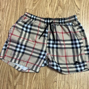 Rutig badshorts från Burberry - Snygga badshorts från Burberry i klassiskt rutigt mönster med beige, svart och röd färg. De har en elastisk midja med snörning för perfekt passform. Perfekta för sommarens alla badäventyr!