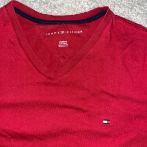 Röd t-shirt från Tommy Hilfiger - Snygg röd t-shirt från Tommy Hilfiger med v-ringning och det klassiska logotypmärket på bröstet. Perfekt för en avslappnad stil. Stor i storleken