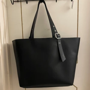 Svart shoppingväska från Even&Odd - Säljer en stilren svart shoppingväska från Even&Odd. Väskan är rymlig och perfekt för både vardag och shoppingturer. Den har en enkel design med ett justerbart band och är tillverkad i syntetmaterial.