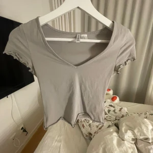 Grå topp - Säljer en grå v-ringad topp från H&M . Toppen har korta ärmar med volangdetaljer och är tillverkad i ett mjukt material. Aldrig använd 