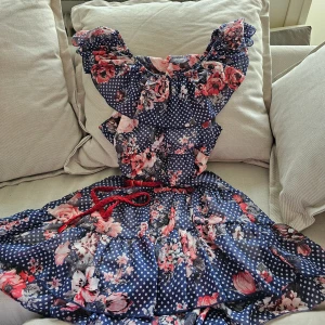 Blommig klänning med volangdetaljer - Charmig klänning i blått med vita prickar och blommönster i rosa och rött. Klänningen har volangdetaljer och ett rött knytband i midjan. Perfekt för sommarens festligheter.Har foder ! 