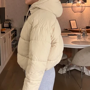 Beige pufferjacka från Monki - Säljer en snygg beige pufferjacka från Monki. Jackan har en croppad design med huva och långa ärmar. Perfekt för kyliga dagar när du vill hålla stilen.