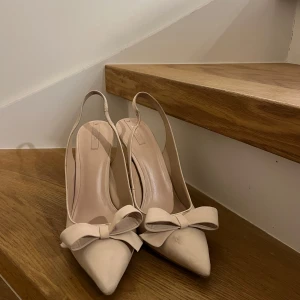 Beige pumps med rosett - Eleganta beige pumps med spetsig tå och en dekorativ rosett framtill. Perfekta för en stilren look.