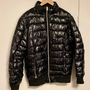 Svart pufferjacka från Smog - Snygg svart pufferjacka från Smog med glansig yta och dragkedja framtill. Jackan har långa ärmar och ribbade muddar vid ärmslut och nederkant. Perfekt för kyligare dagar. Litet hål förekommer i höger nederkant men syns knappt.