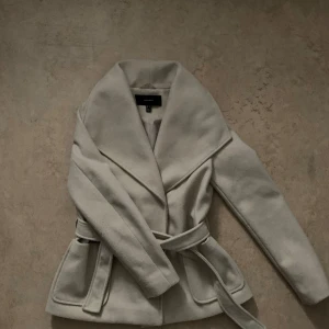 Beige kappa från Vero Moda - Säljer en superfin beige kappa från Vero Moda i storlek S. Den har en elegant design med ett bälte i midjan och stora fickor fram. Perfekt för höst och vinter 😊