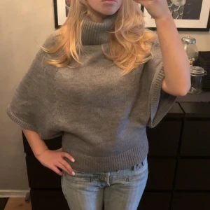 Grå stickad tröja - Säljer en supermysig grå stickad poncho med polokrage. Perfekt för höst och vinter när du vill hålla dig varm och stilren. Den har en loose passform och design som gör den lätt att bära över andra plagg. Perfekt för en avslappnad men chic look!